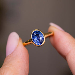 A close-up of a yellow gold Vintage-inspired solitaire engagement ring with an oval blue Sapphire held between two fingers against a soft grey background.