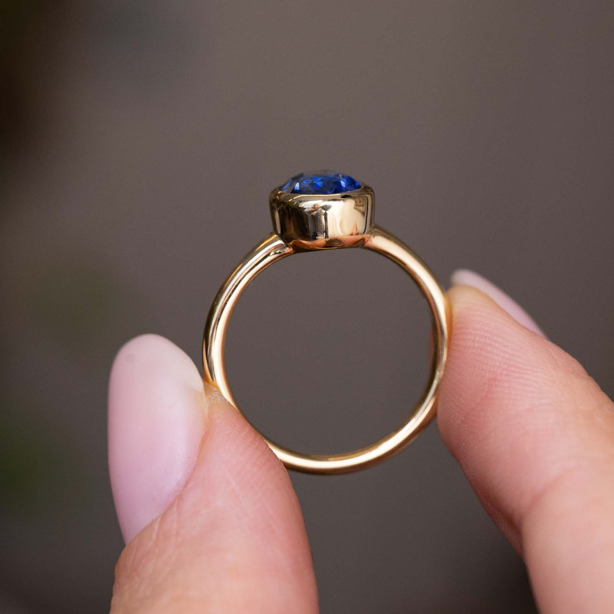 A close up of the side profile of an Antique-inspired, Blue sapphire solitaire engagement ring, held between two fingers against a soft, blurred gray background.  Showcasing the yellow gold band and rub-over setting of the sapphire. 