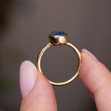 A close up of the side profile of an Antique-inspired, Blue sapphire solitaire engagement ring, held between two fingers against a soft, blurred gray background.  Showcasing the yellow gold band and rub-over setting of the sapphire. 