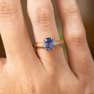 A hand up close wearing a Vintage-inspired, Blue sapphire and diamond flanked solitaire engagement ring in yellow gold.