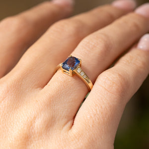 A yellow gold Vintage-inspired, Blue sapphire and diamond flanked solitaire engagement ring worn on the hand and shown from a low close angle, allowing you to see the pierced gallery.