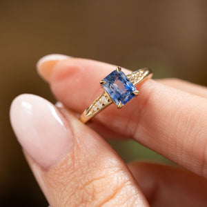 A close-up of a hand wearing a yellow gold Vintage-inspired, Blue sapphire and diamond flanked solitaire engagement ring set against a soft, blurred brown background.