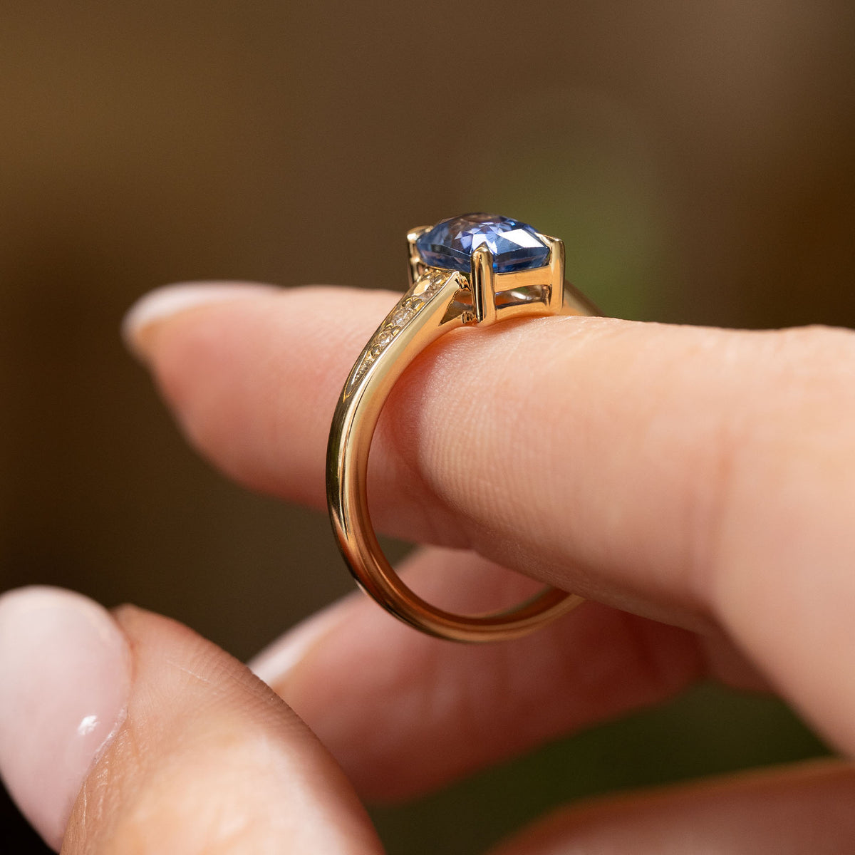 A close image of a finger that is balancing a yellow gold engagement ring set with a natural blue Sapphire and diamond set shoulders. The ring is shown completely from the side showcasing its profile and shank. set against a soft, blurred brown background.
