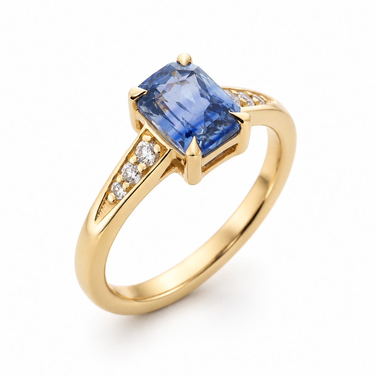 A three-quarter view of a vintage-inspired yellow gold engagement ring set to centre an emerald-cut blue sapphire flanked by diamond, in 14ct yellow gold. 