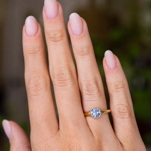 A close-up of a hand wearing a yellow gold Vintage-inspired engagement ring with a square-cut blue sapphire from Sri Lanka, set against a blurred green and brown background.