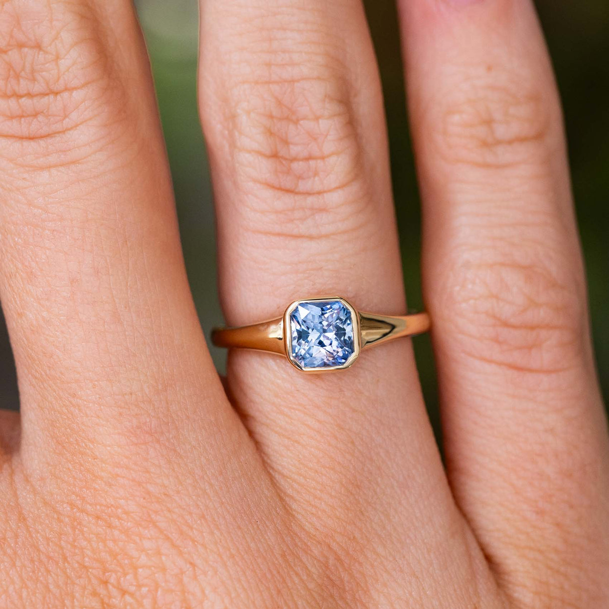 A close-up of a hand wearing a Vintage-inspired, blue square-cut sapphire solitaire engagement ring with a yellow gold bezel setting and band.