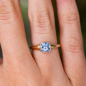 A close-up of a hand wearing a Vintage-inspired, blue square-cut sapphire solitaire engagement ring with a yellow gold bezel setting and band.