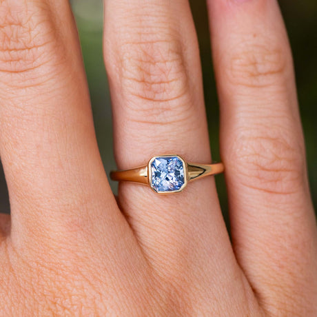 A close-up of a hand wearing a Vintage-inspired, blue square-cut sapphire solitaire engagement ring with a yellow gold bezel setting and band.