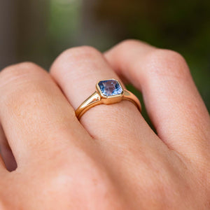 A close-up of a hand wearing a yellow gold solitaire engagement ring with a square-cut light blue sapphire from Sri Lanka, the design is inspired by Antique rings. set against a soft, blurred green background.