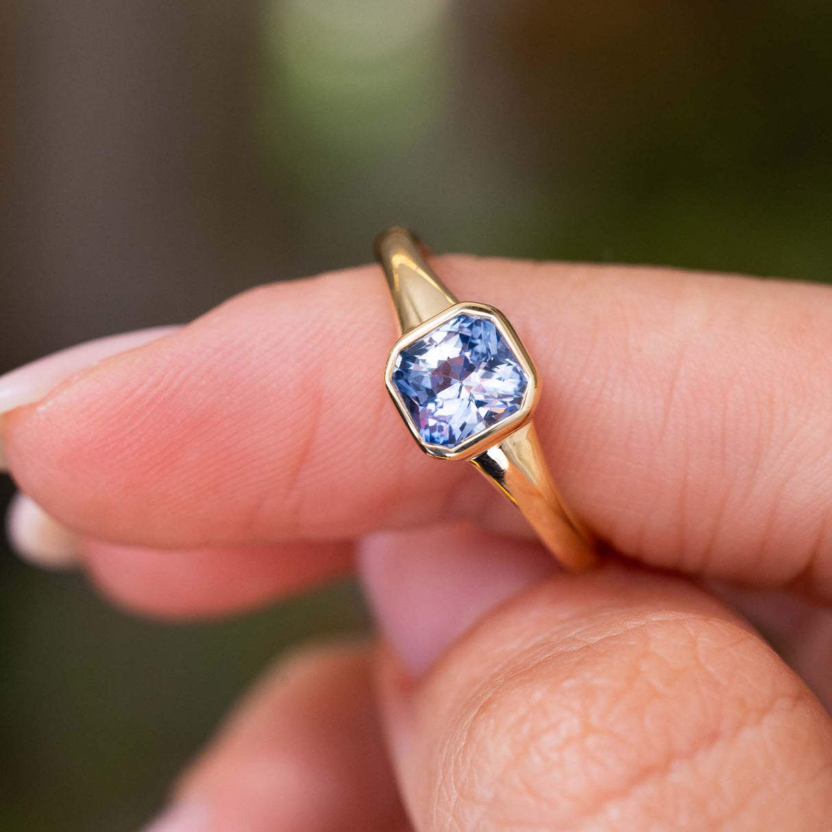 A yellow gold Antique-inspired solitaire engagement ring with a square-cut light blue sapphire and yellow gold setting and band, held between fingers against a blurred green background.
