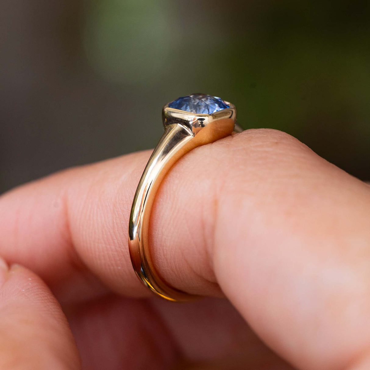 A close up of the side profile and bezel set blue sapphire of an Antique-inspired, solitaire engagement ring in yellow gold. Worn on a finger against a softly blurred background. 