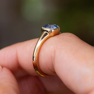 A close up of the side profile and bezel set blue sapphire of an Antique-inspired, solitaire engagement ring in yellow gold. Worn on a finger against a softly blurred background. 