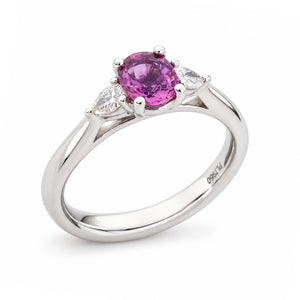 Angled product view of an antique-inspired sapphire ring, featuring a vivid purple-pink centre stone flanked by two pear-shaped diamonds.