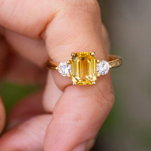 An Antique-inspired, Yellow sapphire and diamond three-stone engagement ring in yellow gold held in a person's finger against a blurry background
