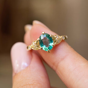 A close-up of a Vintage-inspired, Emerald and diamond engagement ring in yellow gold ring held between fingers against a soft, blurred brown background.