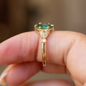 A close-up of a gold ring with a green emerald trilogy engagement ring, worn on a person's finger against a blurred background.