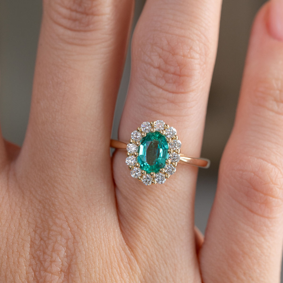 Close finger detail highlighting the richly coloured emerald, sparkling diamond halo and warm yellow gold setting.