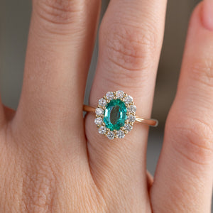 Close finger detail highlighting the richly coloured emerald, sparkling diamond halo and warm yellow gold setting.