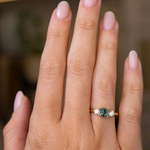 A hand wearing an Antique-inspired, Teal sapphire and diamond three-stone engagement ring in yellow gold.