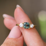 A close-up of a yellow gold Antique-inspired, Teal sapphire and diamond three-stone engagement ring balanced on the fingers and against a blurred background