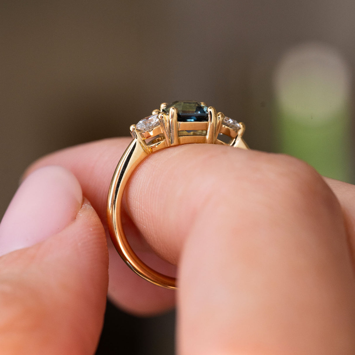 A close-up of a yellow gold ring being held between fingers, Antique-inspired, Teal sapphire and diamond three-stone engagement ring shown from the side profile.