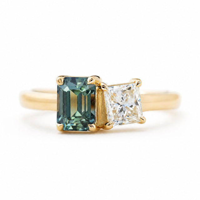 A yellow gold ring featuring an emerald-cut green gemstone and a princess-cut diamond, against a plain white background.