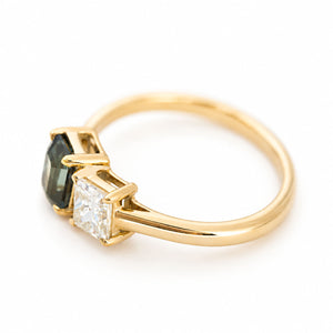 A yellow gold ring featuring teal sapphire gemstone and a princess-cut diamond, set on a clean white background, at an angle.