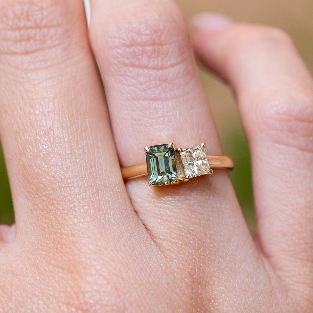 A close-up of a hand wearing a gold ring with an emerald-cut green sapphire and a smaller diamond.