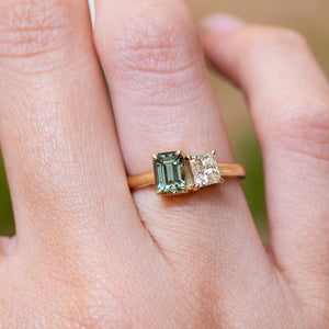 A close-up of a hand wearing a gold ring with an emerald-cut green sapphire and a smaller diamond.