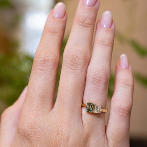 A hand wearing a gold ring with a green rectangular emerald-cut gemstone and a smaller clear stone beside it, against a softly blurred background.