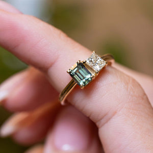 A close-up of a gold ring worn on a finger, featuring a baguette-cut emerald green sapphire and a round princess-cut diamond.