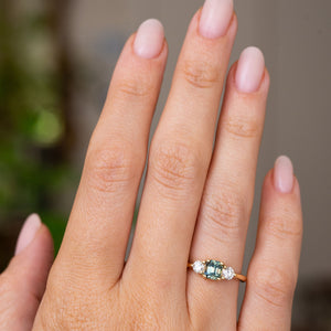 A hand wearing an Antique-inspired, Teal sapphire and diamond three-stone engagement ring in yellow gold to show scale.