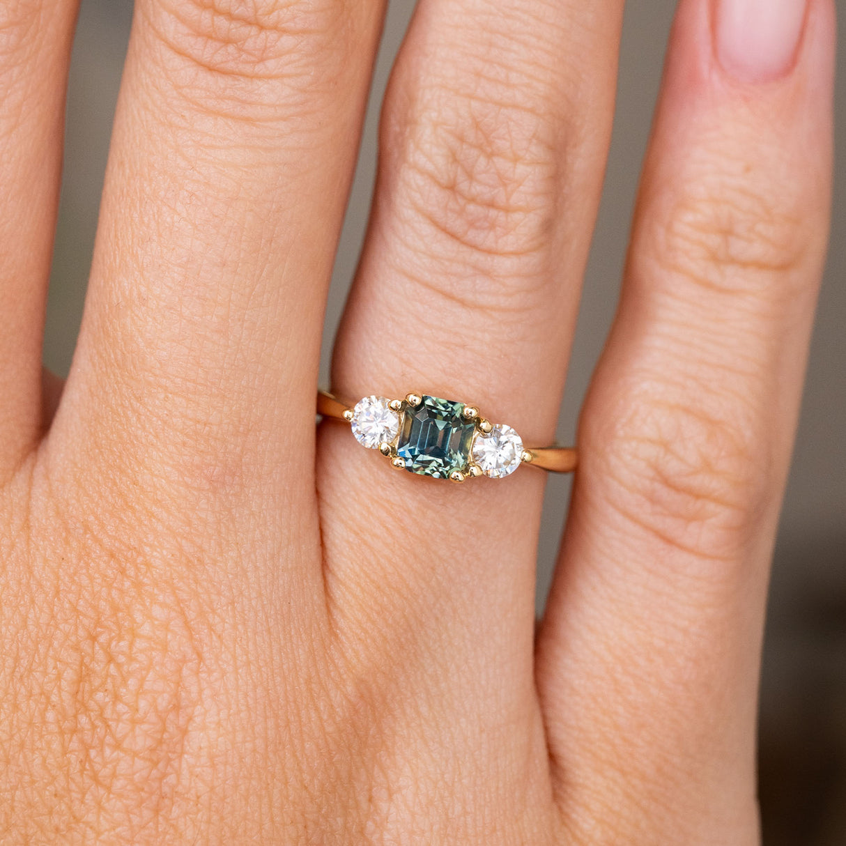 A detailed image of an Antique-inspired, Teal sapphire and diamond three-stone engagement ring in yellow gold worn on a hand
