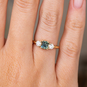A detailed image of an Antique-inspired, Teal sapphire and diamond three-stone engagement ring in yellow gold worn on a hand