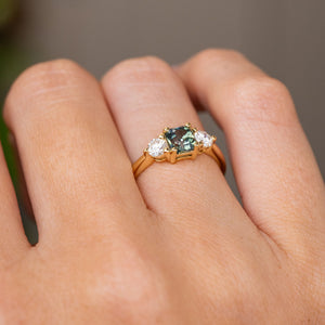 A close up image of an Antique-inspired, Teal sapphire and diamond three-stone engagement ring worn on a finger and shown from a low angle to display the gallery.
