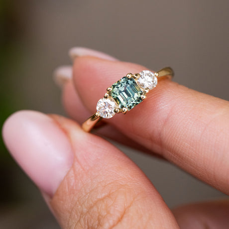 A close-up of a yellow gold Antique-inspired, Teal sapphire and diamond three-stone engagement ring worn on a finger with manicured nails.