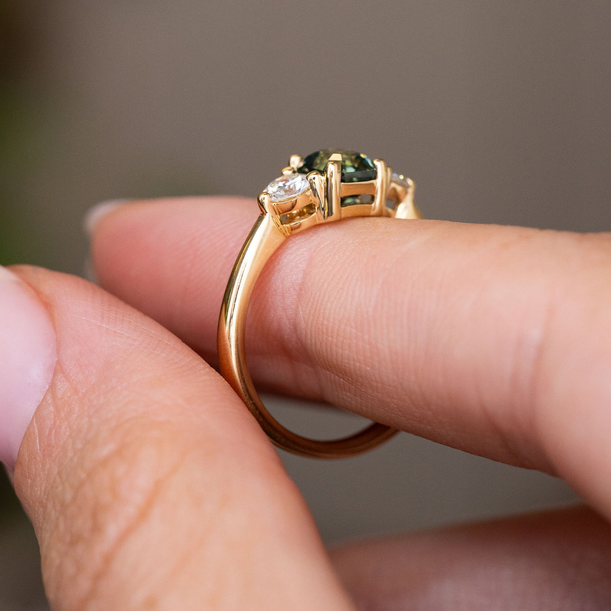 A close-up of a yellow gold Antique-inspired, Teal sapphire and diamond three-stone engagement ring worn on a person's finger against a soft blurred background.