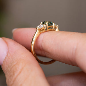 A close-up of a yellow gold Antique-inspired, Teal sapphire and diamond three-stone engagement ring worn on a person's finger against a soft blurred background.