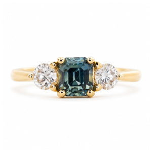 An antique-inspired engagement ring with a teal sapphire at the centre with a round brilliant-cut diamond on each side.