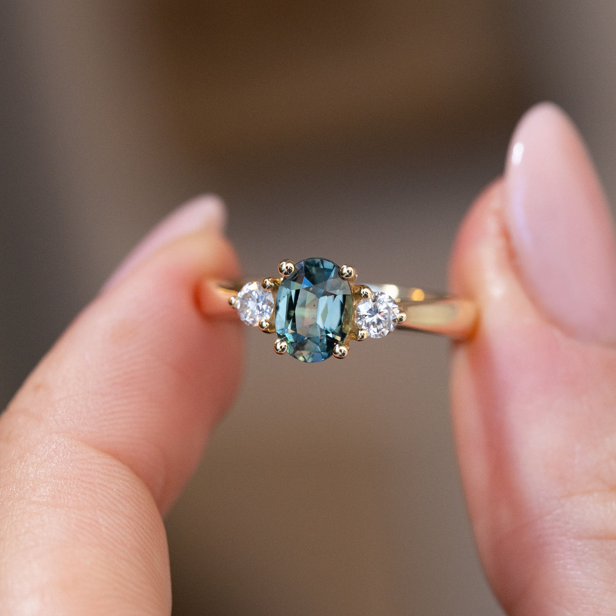 Hand-held close-up of the blue-green oval sapphire and brilliant-cut diamond shoulders in polished yellow gold.