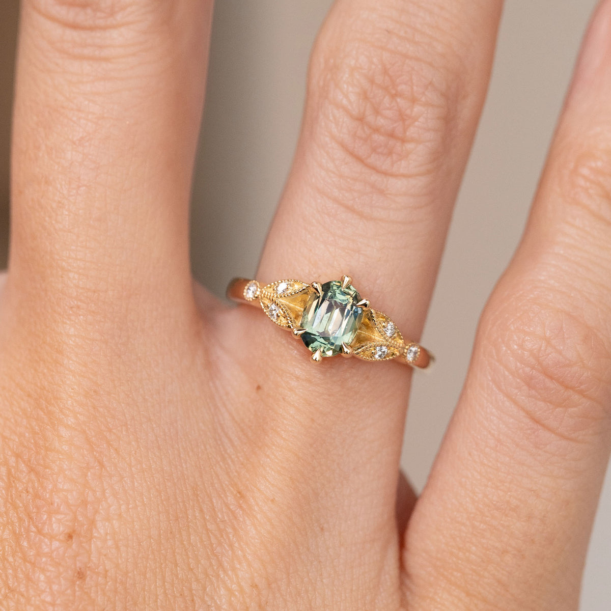 Close-up of the yellow gold engagement ring, with an oval teal sapphire framed by intricate diamond-set shoulders.
