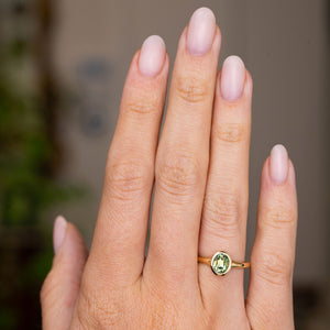 A close-up of a hand wearing a yellow gold Antique-inspired ring on a wedding finger.  An oval green Sri Lankan sapphire is set in the yellow gold band, 