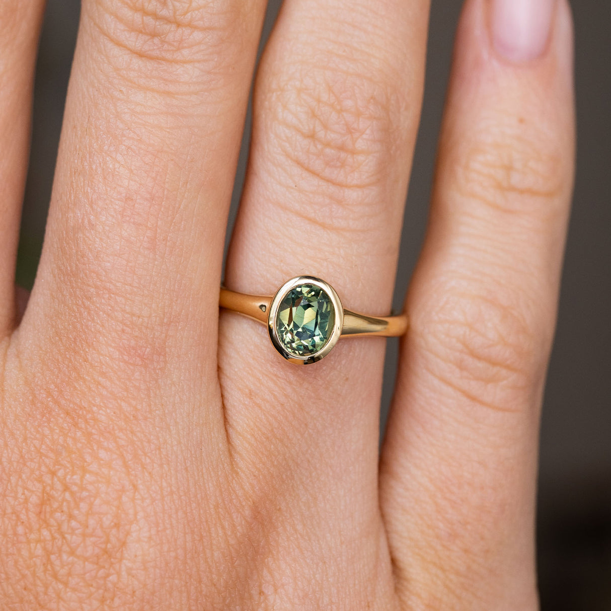 An Antique-inspired, oval green sapphire solitaire engagement ring with a yellow gold bezel setting and band worn on a person's finger, shown in a close-up shot.