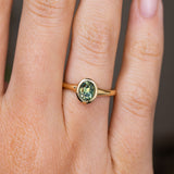 An Antique-inspired, oval green sapphire solitaire engagement ring with a yellow gold bezel setting and band worn on a person's finger, shown in a close-up shot.