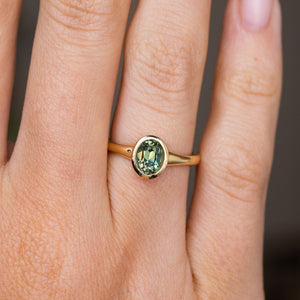 An Antique-inspired, oval green sapphire solitaire engagement ring with a yellow gold bezel setting and band worn on a person's finger, shown in a close-up shot.