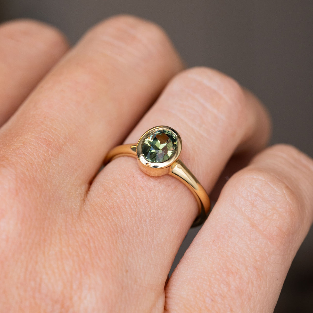 A close up of a yellow gold solitaire engagement ring, with the focus on the oval green Sri Lankan sapphire that is set in an antique-inspired bezel setting worn on a person's finger against a soft grey background.