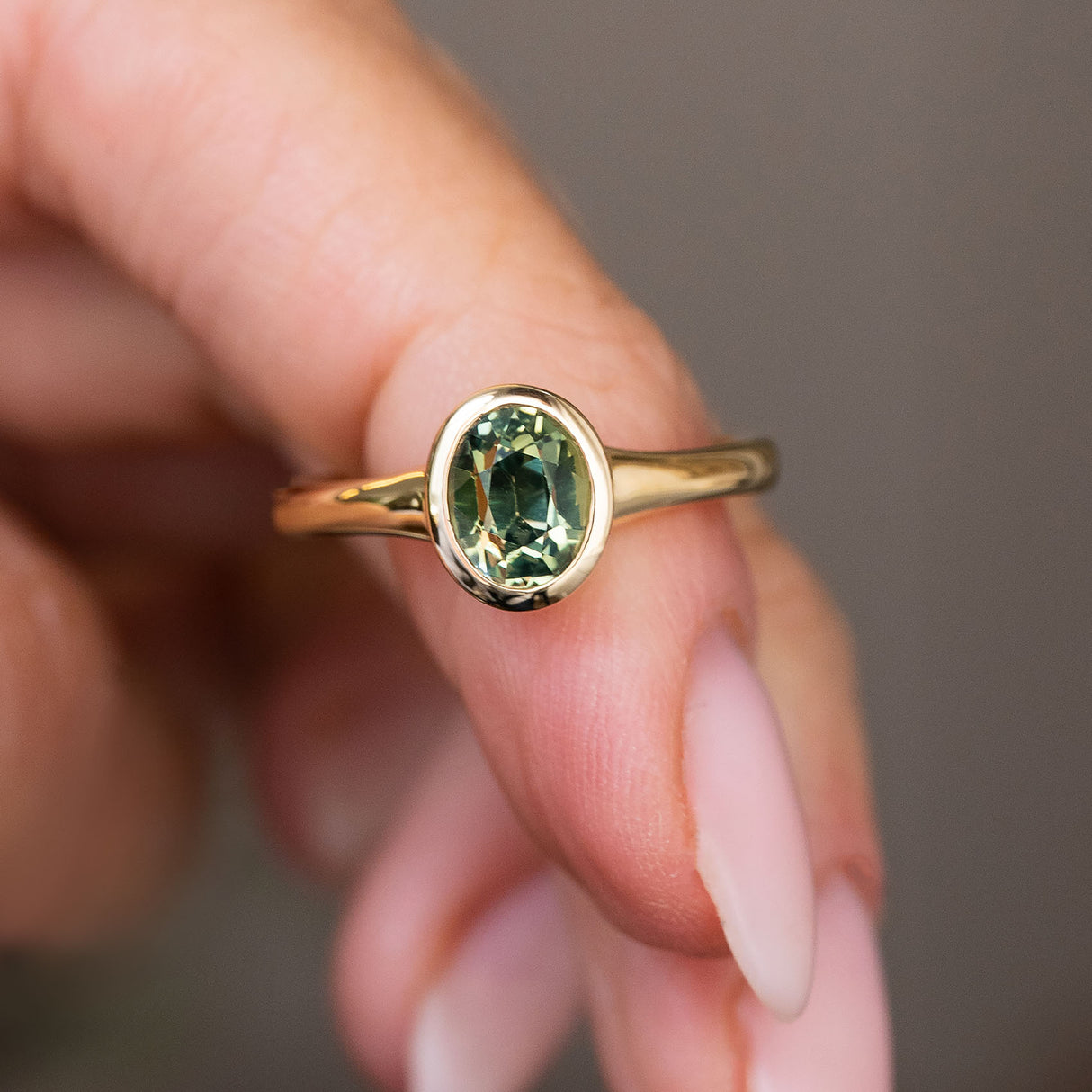 An Antique-inspired, Green sapphire solitaire engagement ring in yellow gold held on a person's finger against a soft grey background.