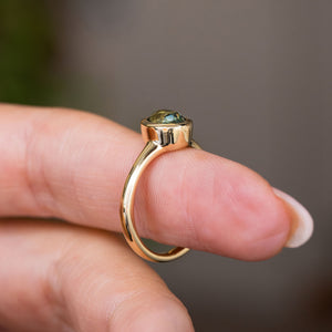 A Vintage-inspired yellow gold solitaire engagement ring set with an oval green sapphire, shown worn on a person's finger against a soft blurred grey background.