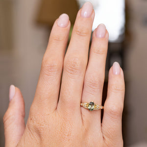 A close-up of a hand wearing a Vintage-inspired, Green sapphire and diamond engagement ring set against a softly blurred indoor background.