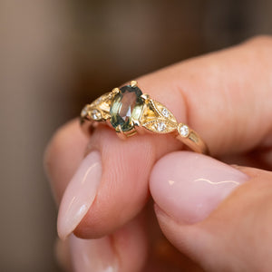 A close-up of a hand holding a gold ring featuring a Vintage-inspired, Green sapphire and diamond engagement ring on a soft blurred background.